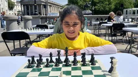 BBC A young girl in a yellow t-shirt sits at a chess board set up on a table in Trafalgar Square in central London. She is facing the camera and is in the middle of a game. There are other tables set up for a chess game in the background.