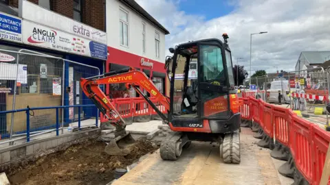 JOANNA TAYLOR/BBC A digger scooping up dirt in front of new paving slabs. To the left, there are shops including Chocoberry, which has a red store front.