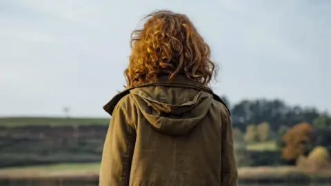 Getty Images A young person with curly hair faces away from the camera, looking at a green landscape. They are wearing a hooded green jacket. 