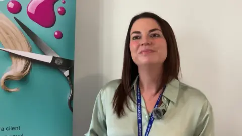 A woman with dark hair and dark eyes wears a green silky shirt and lanyard blue college landyard. She is staning next to a sign which shows scissors and a lock of blonde hair and splashes of pink nail varnish
