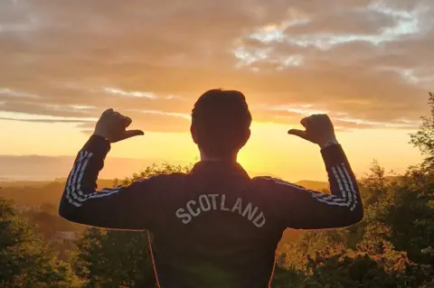 Chris Clark A man wearing a top which says SCOTLAND on the back, with his back to the camera, and a sunrise in the distance.
