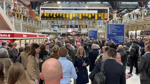 A very busy concourse at London Liverpool Station, under a glazed roof. There are crowds of people in the foreground and stretching back towards the large electronic train information sign, which has bright yellow lettering on it, with London Liverpool Street written across its top. 