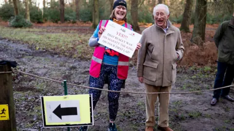 Aaron Chown/PA A woman wearing a pink hi-vis jacket holds a sign that says "YES!!! THAT REALLY WAS HIS MAJESTY THE KING ON YOUR FIRST LAP!" King Charles III is standing next to her in a brown coat and trousers. There is woodland behind them.