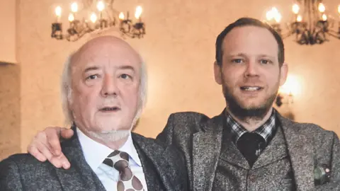 Adam Riding John and Adam Riding poses together in smart grey suits and ties in a grand room with candle-esque lights