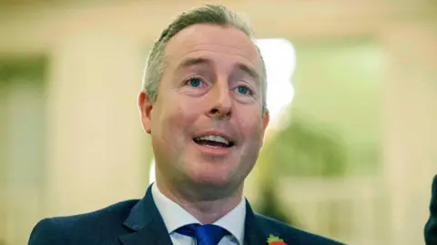 PA Media Paul Givan wearing a navy blazer, white shirt and blue tie. He has short brown hair. The background is blurry. 