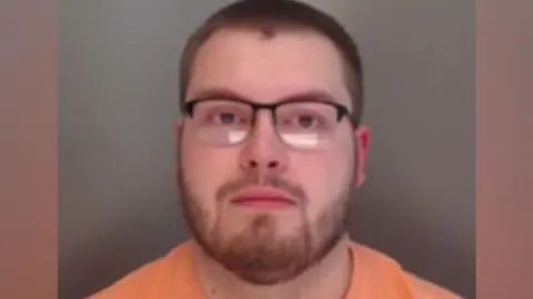 A mugshot of Sains looking into the camera. He has short, brown hair and a short beard. He is wearing rectangular glasses and an orange t-shirt.