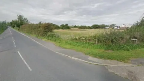 A road running alongside a field with a series of buildings in the distance.