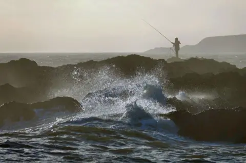 Colin Denholm Huge waves in the foreground in front of a rocky outcrop which a man is standing on makes it look like he is fishing in a treacherous situation.