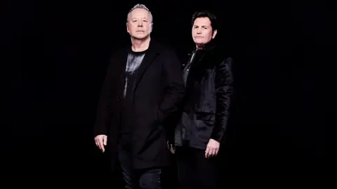 Cuff & Taylor Two men, standing with a black background, looking at the camera. Jim Kerr, is wearing black, with short grey hair, and Charlie Burchill, is also wearing all black. 