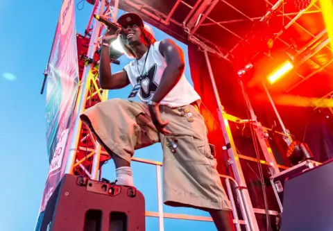 Paul Newbon Photography A performer stands on an outdoor festival stage, holding a microphone and leaning forward with one foot raised on a speaker. Bright red and orange stage lights surround the scene, with metal rigging and banners visible against a blue sky.