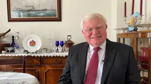Dr Tim Brain wearing a burgundy tie and white shirt, with a black suit jacket. He is sitting in what looks like a dining room with a covered table to his right and some trinkets on a shelf behind him. There is a cabinet with bits in behind him to his left. He is smiling at the camera, wearing glasses, with his grey hair neatly combed.