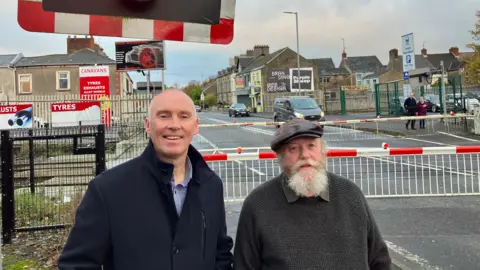 Two men standing in front of a level crossing with the barrier down.