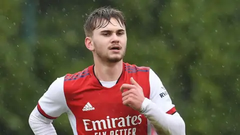 A young man with blonde hair wearing a red and white Arsenal football shirt while running in the run
