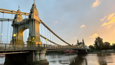The illuminated green and gold tower of Hammersmith Bridge over the River Thames at sunset, with an orange-streaked sky in the background.