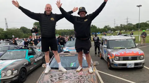 Two men, including Carlos Baldry, dressed in black long sleeved tops and shorts stand on the bonnet of a rust bucket rally vehicle with their arms aloft.
