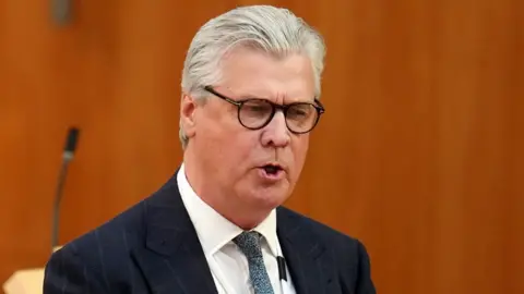Getty Images Malcolm Offord, who has grey hair swept back and black rimmed glasses. He is wearing a business suit with wood panelling behind him 