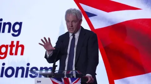 Danny Kruger is on a platform speaking. One had is gesturing as he speaks. A union flag is on a screen behind him. He's wearing a suit.