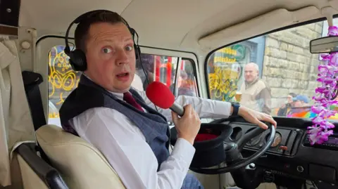 Matt Chorley looks startled as he turns to the camera. He is sitting on the driving seat of a van and holding a red microphone 
