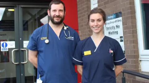 Two medics standing outside the entrance to an emergency department. The man, on the left, has short dark hair and a beard, with a stethoscope around his neck. The woman has dark tied-back hair. Both are wearing dark blue uniform tops and have a relaxed demeanour, with their hands behind their back.
