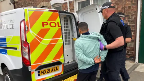 BBC A police van parked outside terraced houses. Two men dressed in all black clothing, with caps on that have police checked patterning on, grab the arm of one smaller man, wearing a light blue tracksuit top and black pants, into the back of the police van.