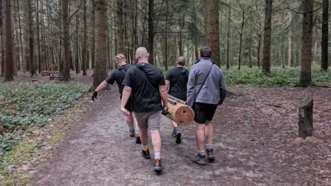 Carrying The Weight Together A group of men carrying a log through the woods