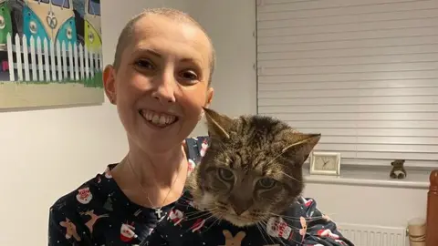 Francesca Vincent is pictured with very short hair. She is smiling at the camera and holding a tabby cat in her arms. They are wearing matching pyjamas with pictures of reindeers and Father Christmas. 