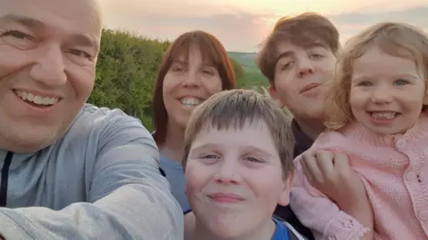 Family handout Three children, two boys and a little girl being held in the air by her brother smile at the camera. Serena is in the middle of the family group with her husband Mark to the side of the image. The whole family is huddled together with a sunset behind them, smiling.