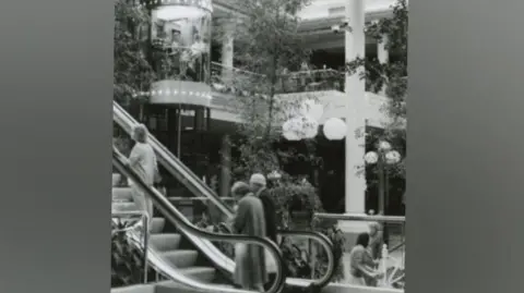 Metrocentre A black and white photo of an older couple going up on an escalator. There is a woman in front of them. To their right there is a tubular lift which has people inside of it. There is a white pillar in the centre of the room which is large and open with a glass ceiling. There are trees decorating the space.