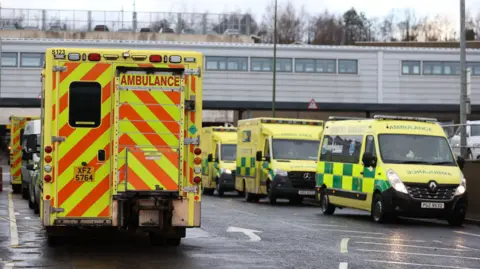 A row of yellow emergency ambulances are lined up outside a grey brick emergency department. 