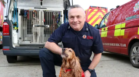 Essex County Fire and Rescue Service Graham Currie kneels behind service dog Jarvis. Mr Currie is wearing a navy fire service uniform while Jarvis is a chestnut coloured dog with a red and orange coloured collar and lead on him. Fire service vehicles are parked behind them.