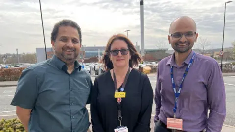 University Hospitals of North Midlands NHS Trust A woman and two men stand together in a hospital car park and smile at the camera. 
