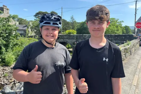 Two teenage boys looking at the camera. One is wearing a bike helmet and a grey top. He is smiling at the camera. The other is wearing a black top with an under armour logo. He is brown hair. 