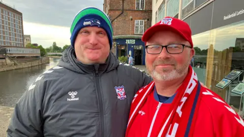 Seb Cheer/BBC Two men standing on a bridge. They are both in football kits and scarves and smiling at the camera