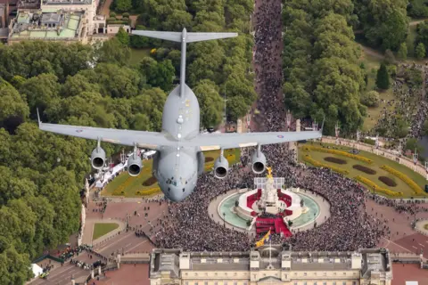 AS1 Amelia Turnbull/PA Wire A grey C17 Globemaster bomber plane is pictured flying through the air, having been captured by a photographer positioned on another aircraft above it which is out-of-frame. Below the aircraft a huge crowd is seen gathered around the statue and fountain in front of Buckingham Palace with green foliage around them.
