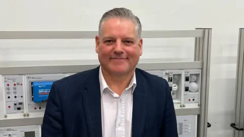 A man with grey hair smiles into the camera - he is wearing a blue checked blazer and white shirt. The man is standing in front of an industrial machine featuring switches and green and red lights.