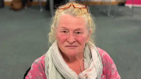 George King/BBC An elderly woman wearing a scarf, pink top and glasses on her head, smiling into the camera 