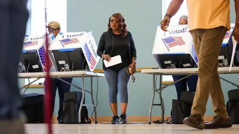 People casts ballots during early voting at the Garners Ferry Adult Activity Center on 26 May 2026 in Hopkins, South Carolina. A woman in a black top and gray leggings walks between two voting tables, holding a white piece of paper and her phone