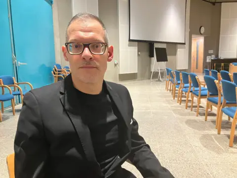 BBC/Amy Holmes Independent Central Bedfordshire councillor Adam Zerny, who has glasses and is wearing a black suit jacket and a black t-shirt. He has short grey hair. He is sitting down. There are rows of blue and brown chairs in the background.