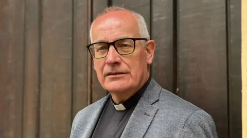BBC/Michelle Lyons A man stands in front of a large dark wooden door or wooden panelled wall. He is positioned slightly to the right of the frame and is looking towards the camera. He is wearing a light grey textured blazer over a dark shirt with a white clerical collar. He has dark rectangular glasses and very short, light-coloured hair around the sides and back.