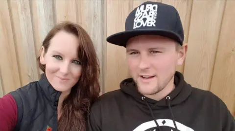 Kirsty Walsh A woman and a man in a selfie. The woman has long brown hair and is wearing a red long-sleeved top under a black gilet. The man is wearing a black hoodie with a large white logo on it, and a black cap with white writing on it