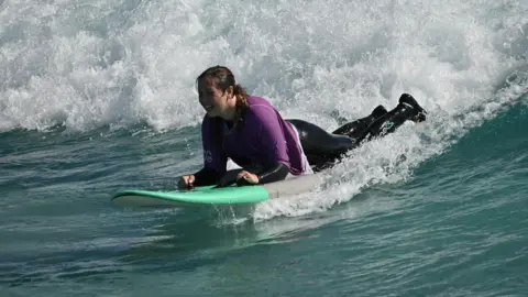 Kay Millar Kay Millar surfing on a wave. She is wearing a purple rash vest on top of a black wetsuit. She is surfing on a green and grey surfboard.