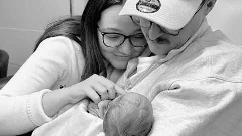 Jade Morris / Joe Bebbington A black and white photo of Jade and Joe holding Felicity., with the top of the baby's head towards the camera. Jade is wearing dark-framed glasses, while Joe has a light-coloured baseball cap on his head