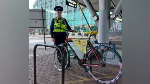 Hertfordshire Police A female police officer stands next to a bike that has brightly coloured wheels with pink and green flecks. There is a yellow sign on the bike with warning messages to thieves and cyclists and the saddle is bright yellow. The bike is chained to a cycle rack outside a shopping centre, with large glass panels that are coloured slightly turquoise. The officer wears a high visibility vest over a blue uniform and a black and blue police hat.