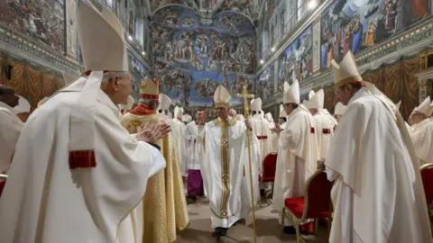 EPA Pope Leo walks down the aisle of the Sistine Chapel. He wears a white robe. Cardinals, also in white robes, applaud as he passes.
