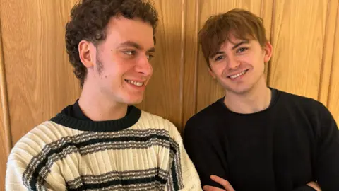 George Usher (Liam Gallagher) standing next to and looking at Oscar Lloyd (Damon Albarn), both standing against a wooden wall with their arms folded