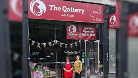 Debbie Morgan Two women stand outside of a cafe with a red sign saying The Qattery. The woman on the left wears a red apron over her shirt while the woman on the right wears a yellow shirt. Progress flags are hung up in the window.