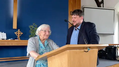 Marjorie Mitchell is standing at a modern wooden lectern with Pastor Sam Davis