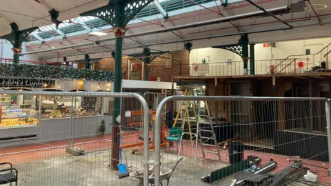 Inside a market hall, there are stalls on the left side and in the middle there is silver protective fencing. Inside the fencing are equipment including a ladder, wheelbarrow and spades