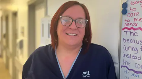 Nurse Katie Anderson smiling at the camera. She's wearing blue scrubs and has short red hair and glasses. She is standing next to a whiteboard with notes. 