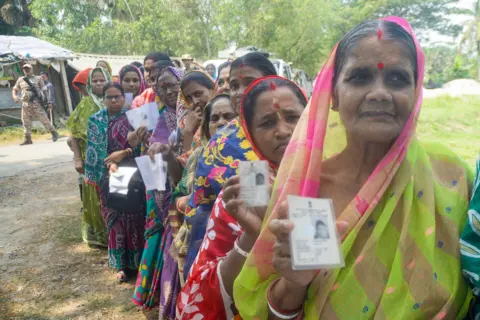 ANI Women in bright green, red and blue saris, red bindis dotting their foreheads, clutch voting cards as they stand in line during West Bengal’s state elections. Behind them, green trees and a tin-roofed hut frame the scene, while a khaki-clad security officer with grey boots and a black rifle keeps watch.